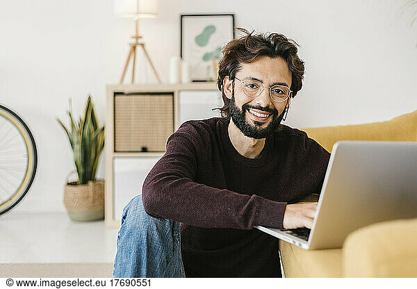 Smiling man with laptop sitting by sofa at home
