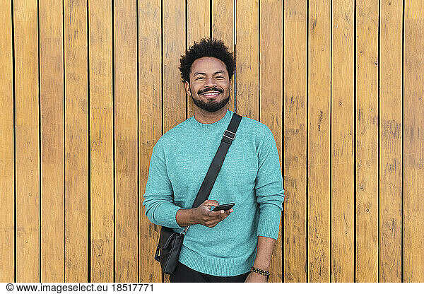 Smiling man with smart phone in front of wooden wall
