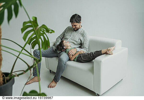 Smiling man with son lying on sofa in front of white wall