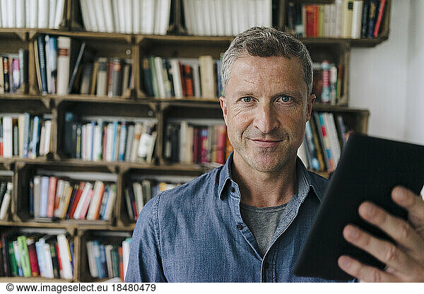 Smiling man with tablet PC standing in front of bookshelf