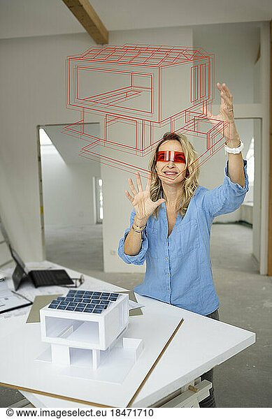 Smiling mature architect wearing virtual reality simulator gesturing and examining house model