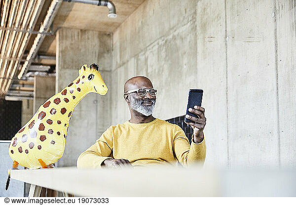 Smiling mature businessman sitting at desk in office with cell phone and giraffe figurine