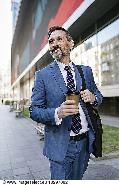 Smiling mature businessman with disposable cup walking on footpath