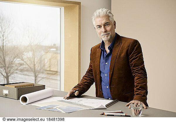 Smiling mature businessman with gray hair standing by table at office