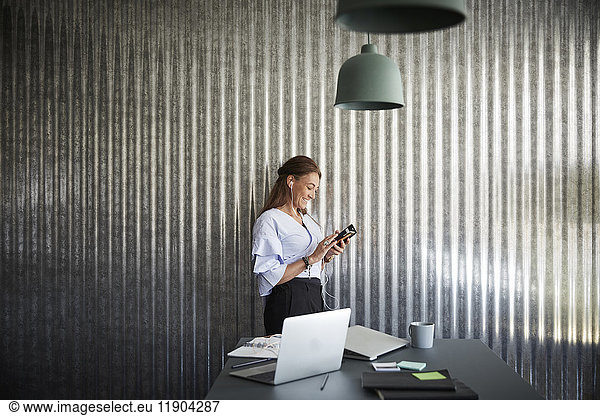 Smiling mature businesswoman using mobile phone while standing against corrugated iron wall at creative office