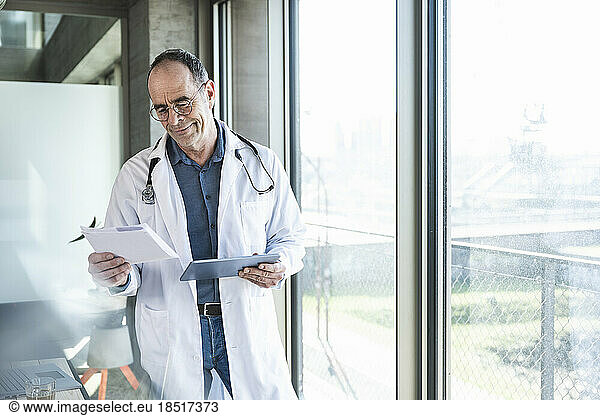 Smiling mature doctor reading prescription and holding tablet PC by window