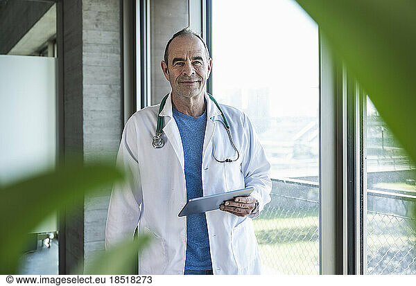 Smiling mature doctor standing with tablet PC by window