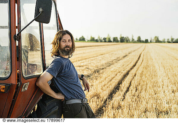Smiling mature farmer leaning on tractor standing at field wheat