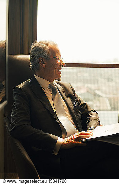 Smiling mature lawyer with documents looking through window at office