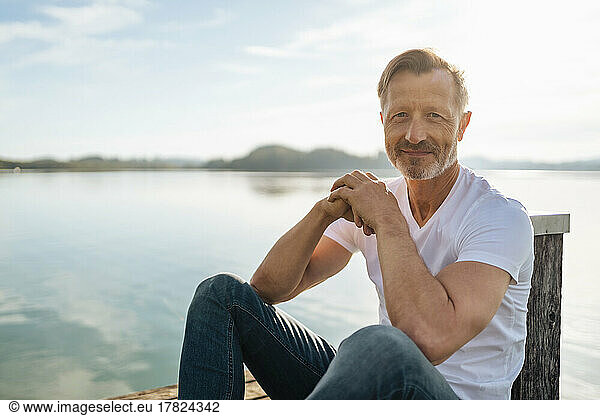 Smiling mature man sitting hands clasped by lake