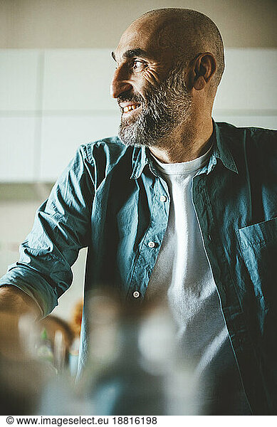 Smiling mature man with shaved head and greying beard sitting in kitchen