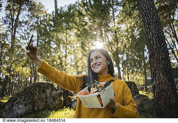 Smiling mature woman holding crate with tangerines taking a selfie
