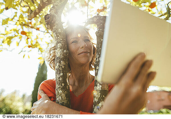 Smiling mature woman hugging orange fruit tree and using tablet PC