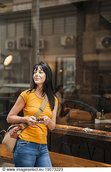 Smiling mature woman leaning on glass with smart phone