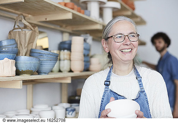 Smiling mature woman looking away while holding bowl in pottery class