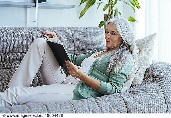 Smiling mature woman reading book on sofa at home