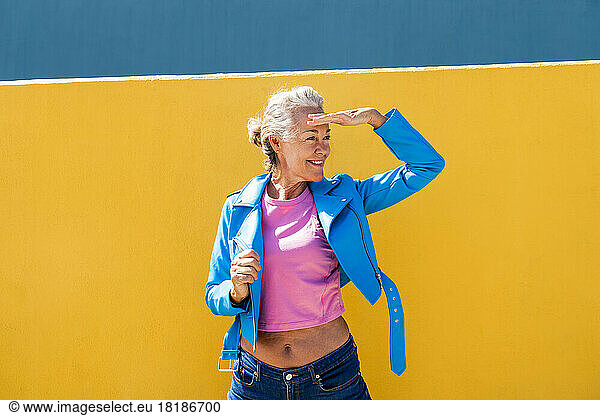 Smiling mature woman shielding eyes in front of colored wall