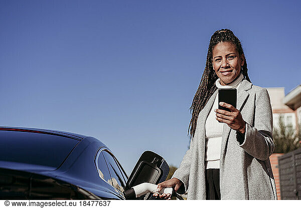 Smiling mature woman standing with smart phone and charging car on sunny day