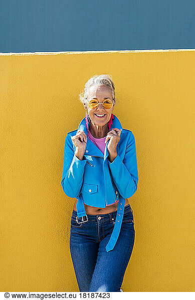Smiling mature woman wearing blue leather jacket leaning on colored wall