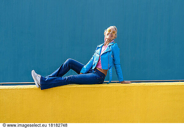 Smiling mature woman wearing blue leather jacket on yellow wall