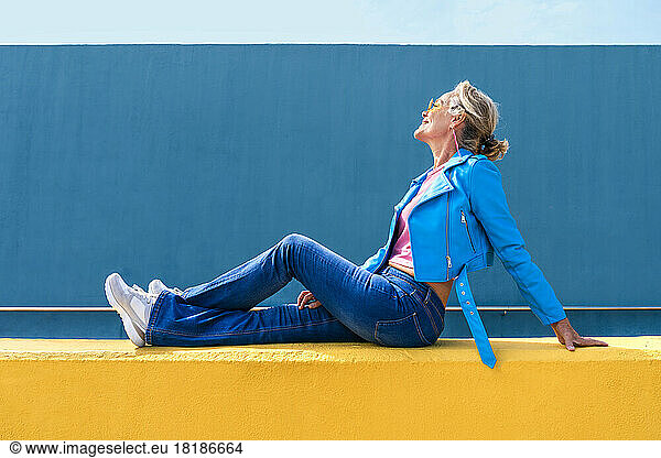 Smiling mature woman wearing blue leather jacket sitting on yellow wall