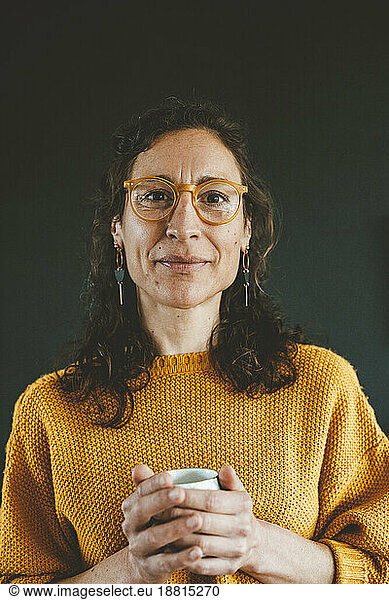 Smiling mature woman wearing eyeglasses holding coffee cup against black background
