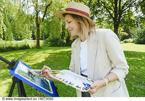 Smiling mature woman wearing hat painting on canvas