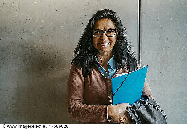 Smiling mature woman with books and file standing against gray wall at university