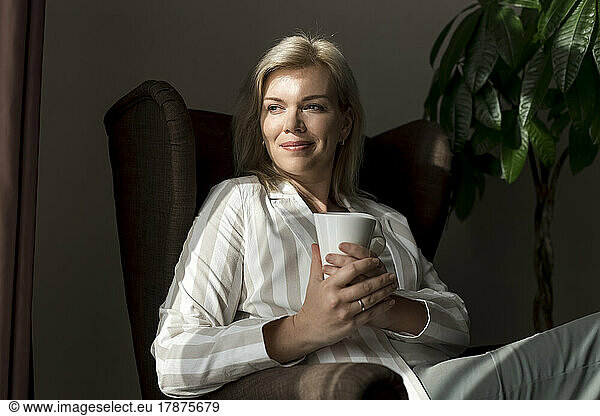 Smiling mature woman with coffee mug sitting in armchair at home