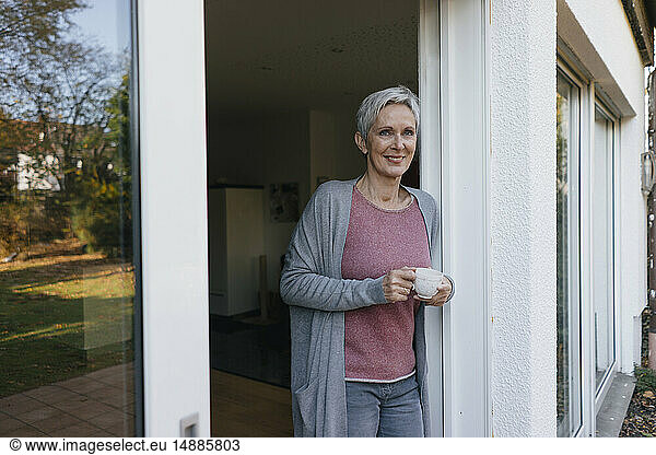 Smiling mature woman with cup of coffee at terrace door