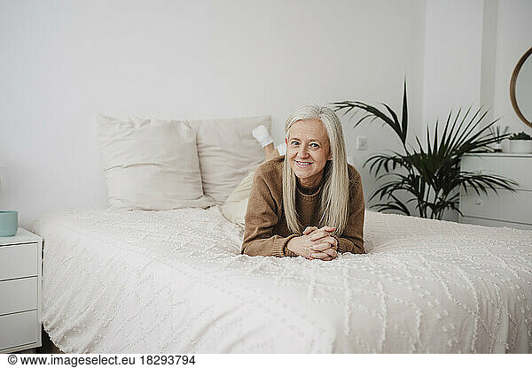 Smiling mature woman with hands clasped lying on bed at home