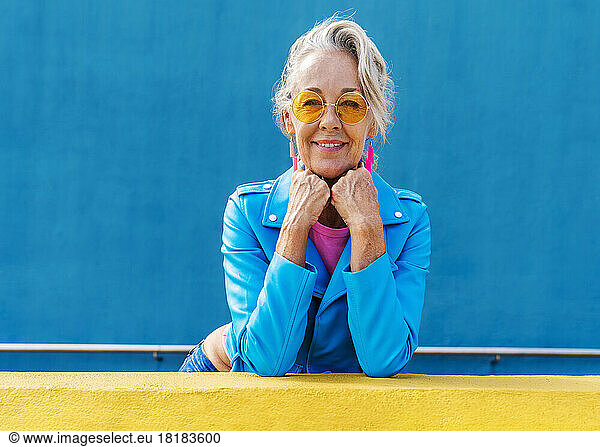 Smiling mature woman with hands on chin in front of blue wall