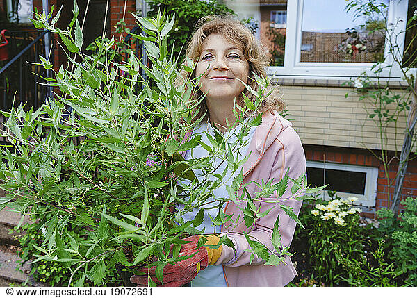 Smiling mature woman with sage plant standing in front of house