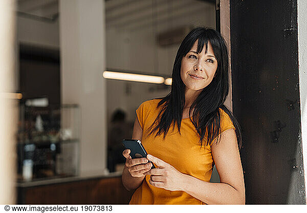 Smiling mature woman with smart phone leaning in doorway