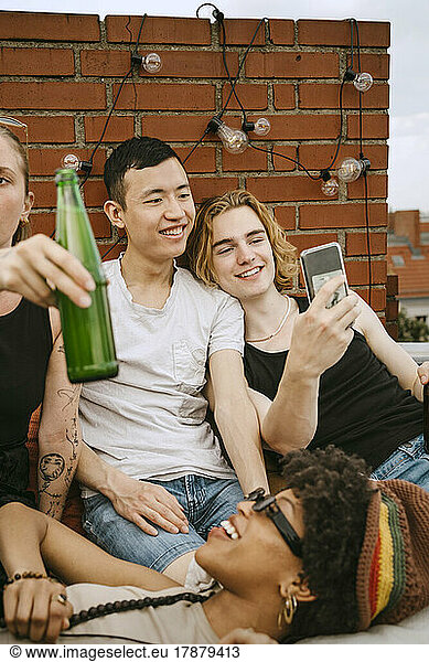 Smiling men sharing mobile phone by female friends on rooftop