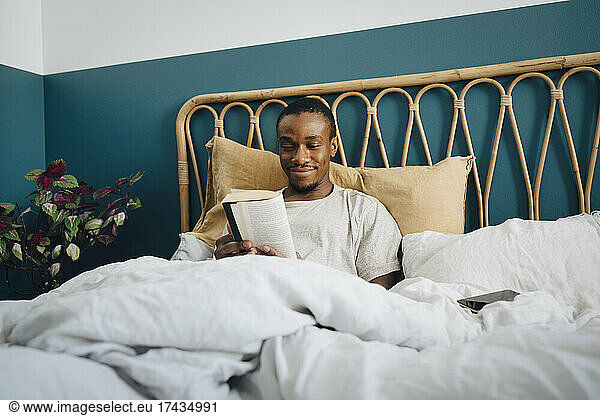 Smiling mid adult man reading book in bed at home