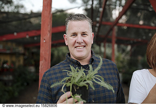 Smiling mid-40's man in blue checked shirt holding plant at nursery