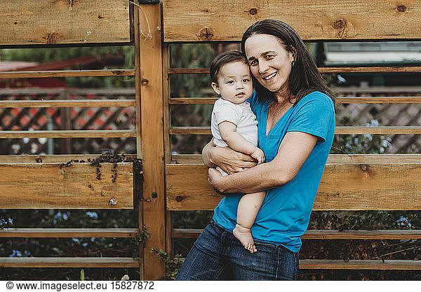 Smiling mid-40's mom holding baby girl outdoors near wooden fence