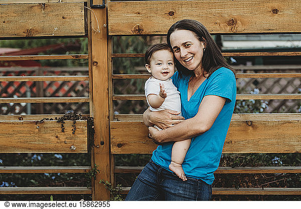 Smiling mid-40's mom holding pudgy baby near lateral wood fence