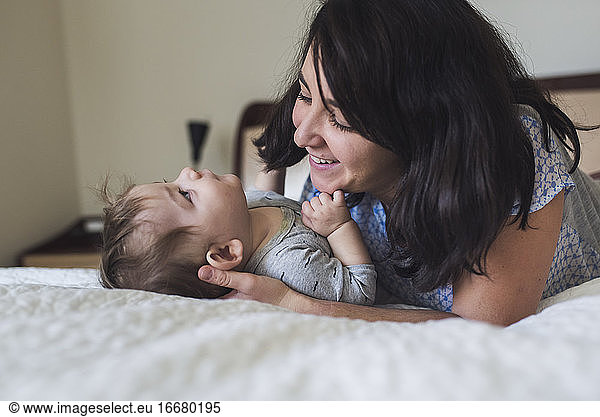 Smiling mid-30's mom with dark hair lovingly admiring baby on bed