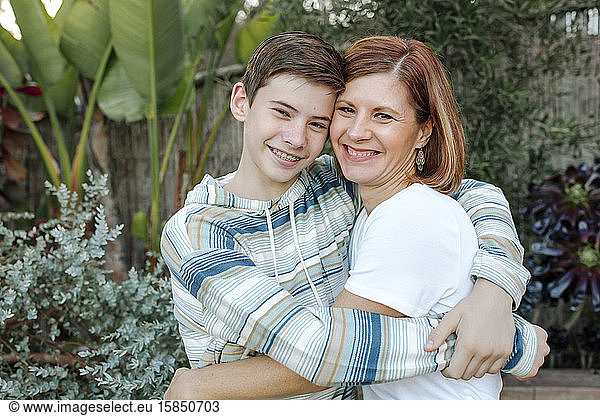 Smiling mom and teen son hugging outdoors with lush plants