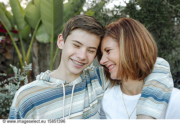 Smiling mom and teen son hugging with large plants in background