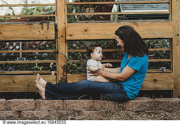 Smiling mom holding baby on outstretched legs in front of fence