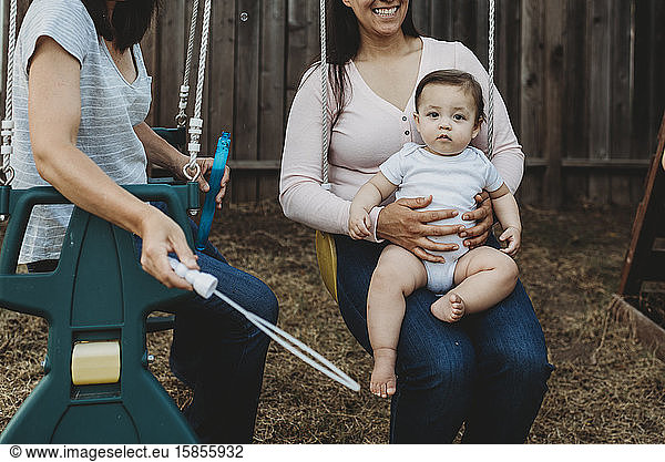Smiling mom holding baby on swing while wife plays with bubbles