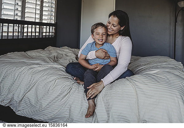 Smiling mom holding happy 5 yr old son on comforter near window