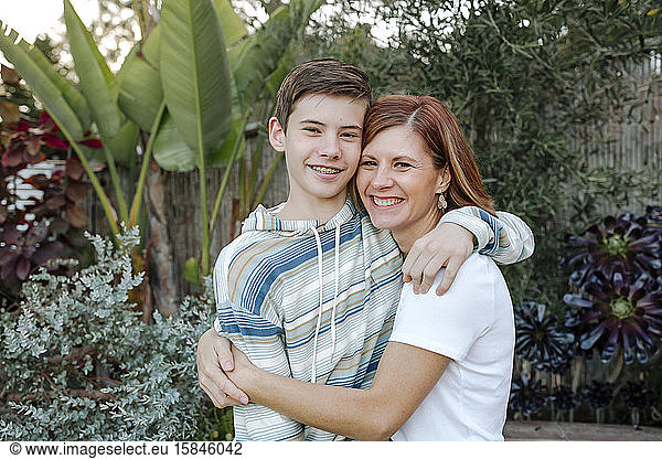 Smiling mom hugging teen son with lush plants on background