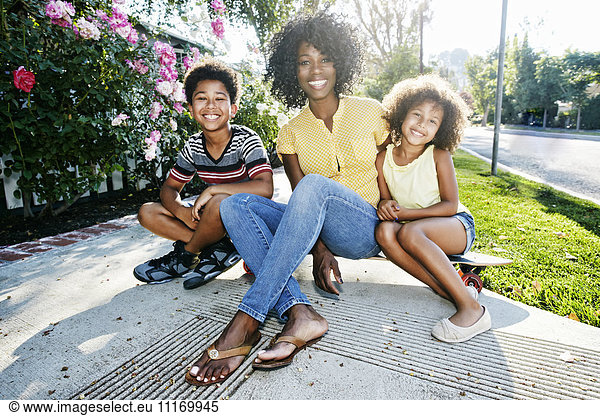 Smiling mother and children sitting on skateboard on sidewalk