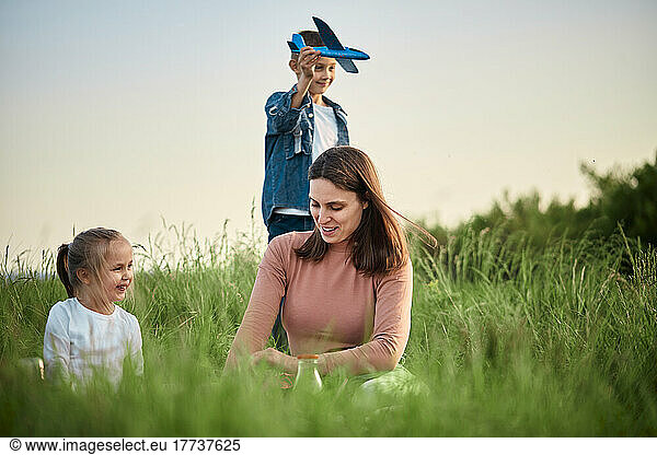 Smiling mother and daughter in front of boy playing with toy airplane on field