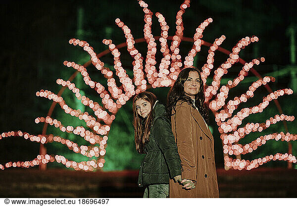 Smiling mother and daughter standing back to back in front of Christmas lights