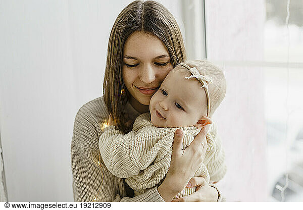 Smiling mother carrying baby daughter near window at home